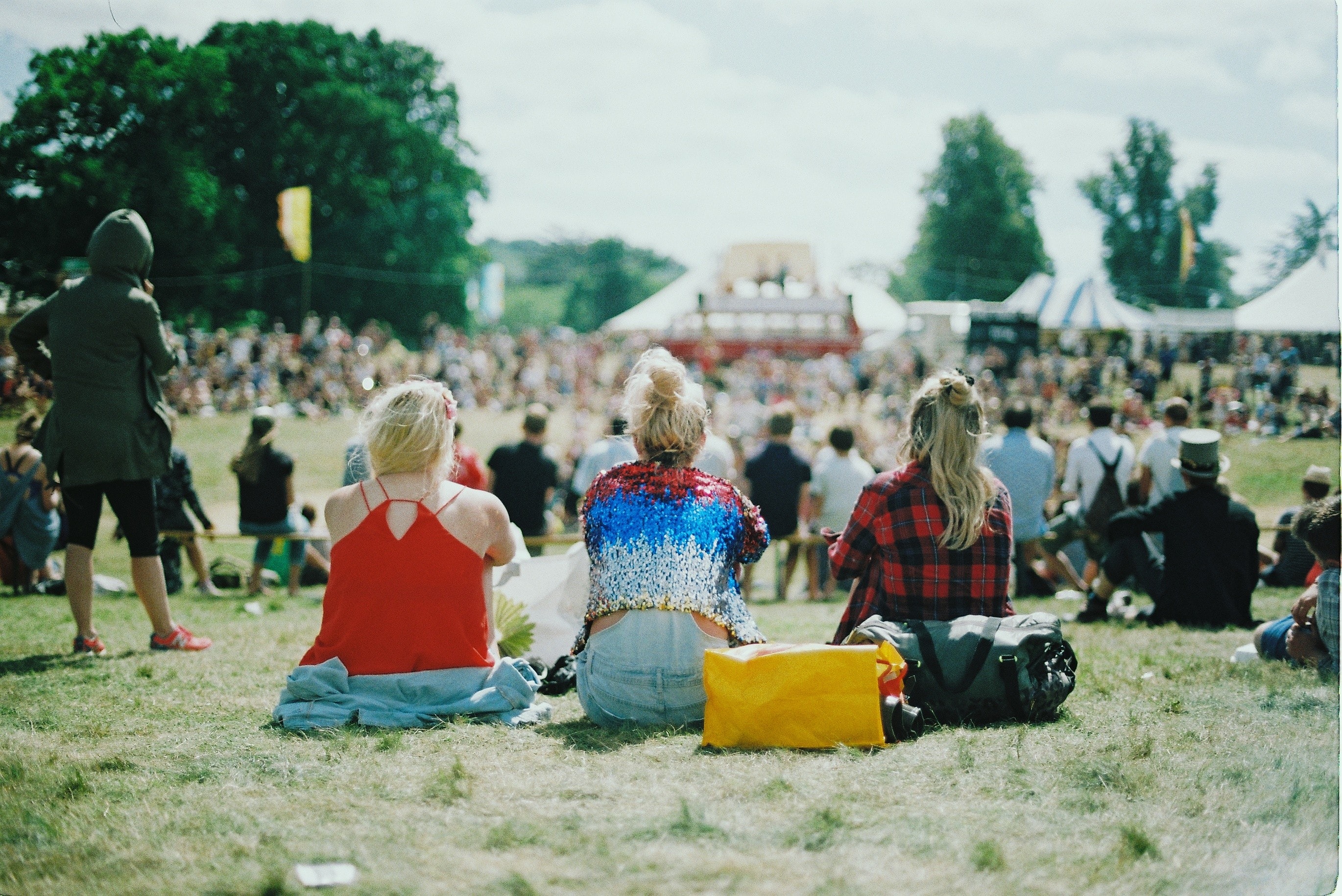 A crowd of people sat on the grass watching music. The focus of the image is on 3 girls in the middle of the photo.