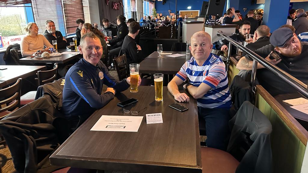 The photo shows two Reading FC fans at a table at The Jazz Cafe drinking beer. They are wearing their Reading FC shirts.