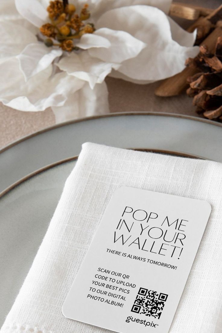 The image shows a table place setting with a white napkin placed on top of a white plate. On top the napkin there is a small piece of paper that reads "Pop me in your wallet" with instructions of how to upload photos from the event to a digital album.