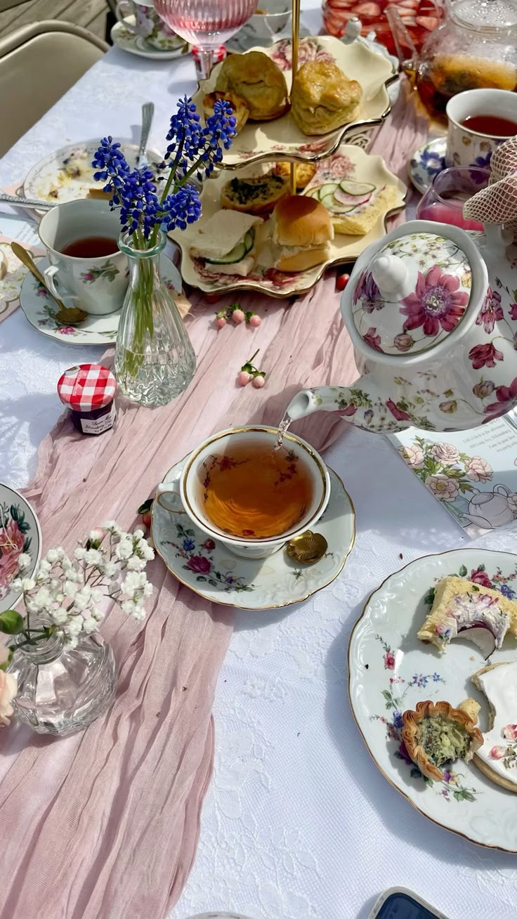 The image shows a table with a pink runner and afternoon tea food placed on it. There is a tea pot that someone is pouring tea into a cup.
