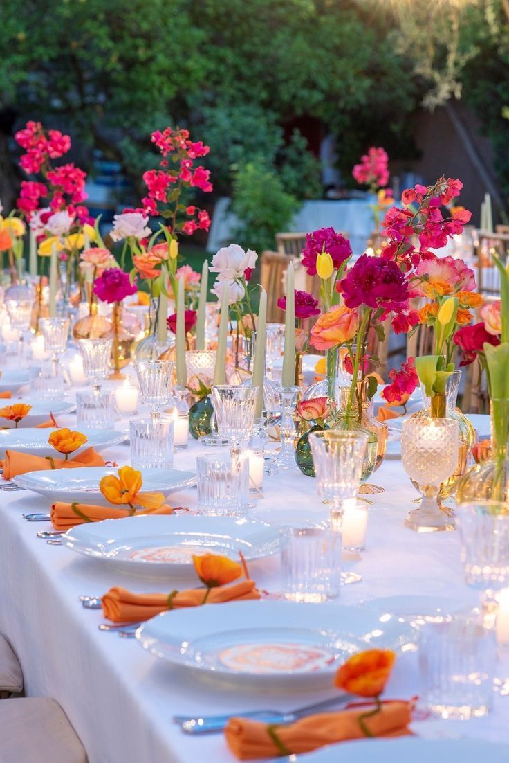 The image shows a table sit outside for dinner. There are bright floral arrangements down the centre of the table with candles and bright orange napkins.