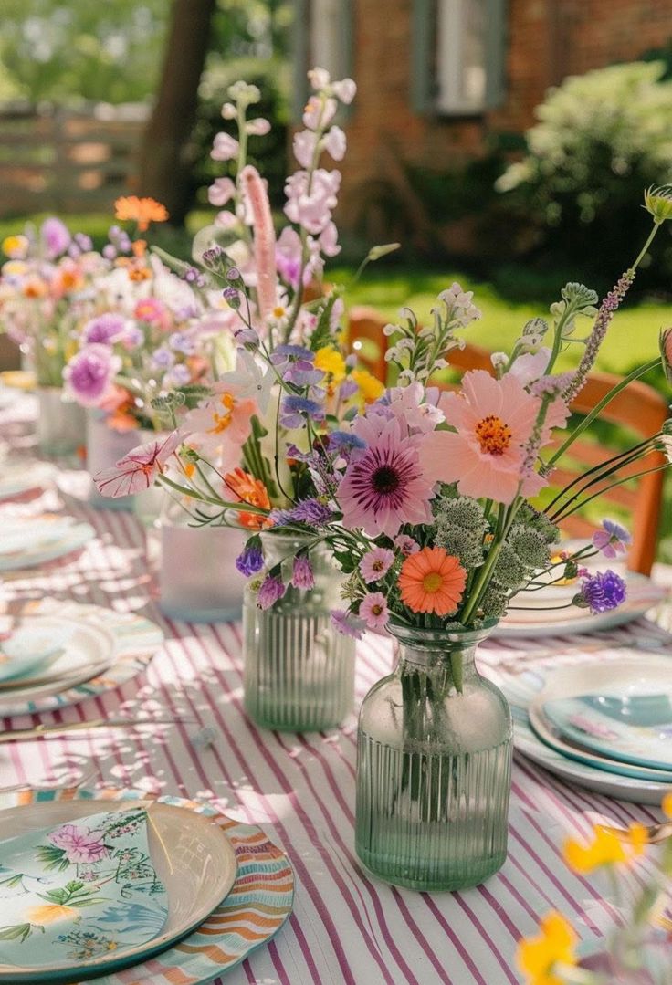 The image shows a table laid for dinner in a garden. There are flowers in vases down the centre of the table.